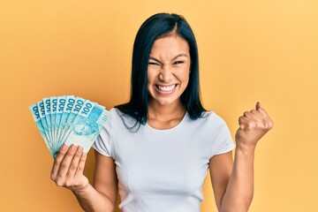 Beautiful hispanic woman holding 100 brazilian real banknotes screaming proud, celebrating victory...
