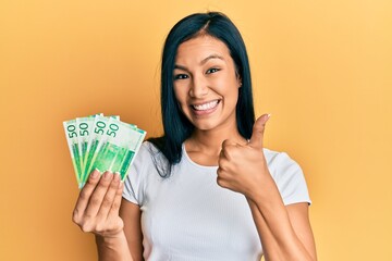 Beautiful hispanic woman holding 50 norwegian krone banknotes smiling happy and positive, thumb up...