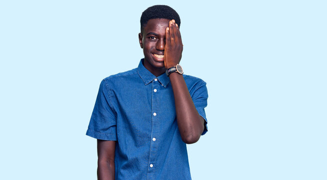 Young african american man wearing casual clothes covering one eye with hand, confident smile on face and surprise emotion.