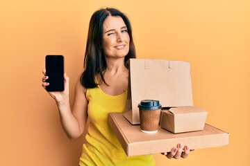 Middle age brunette woman holding take away food showing smartphone screen winking looking at the...