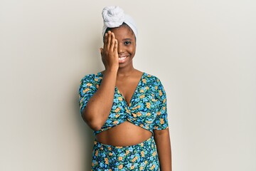 Young african woman wearing hair turban and african style covering one eye with hand, confident smile on face and surprise emotion.