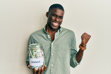 Young african american man holding jar with savings screaming proud, celebrating victory and...