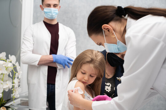 Grandma Cheers Her Granddaughter During The Vaccination. A Young Nurse Stabs A Needle Into The Girl's Arm. A Trainee In The Last Year Of Medicine Observes The Whole Situation In The Doctor's Office