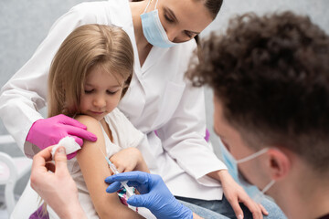 The doctor is getting ready to vaccinate a little girl. A young nurse sits next to the child to...