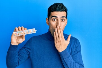Handsome hispanic man holding pill organizer covering mouth with hand, shocked and afraid for...