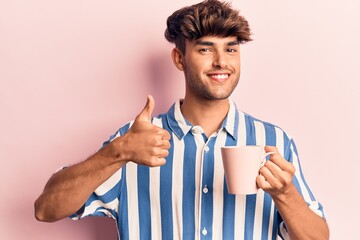 Young hispanic man holding coffee smiling happy and positive, thumb up doing excellent and approval sign