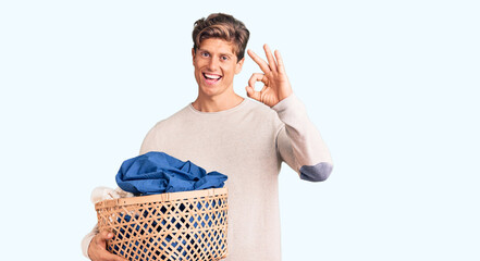 Young handsome man holding laundry basket doing ok sign with fingers, smiling friendly gesturing...