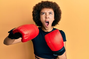 Young hispanic sporty girl using boxing gloves angry and mad screaming frustrated and furious, shouting with anger looking up.