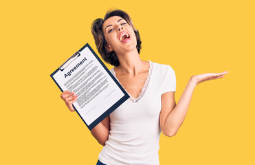 Young beautiful woman holding clipboard with agreement document celebrating victory with happy smile and winner expression with raised hands
