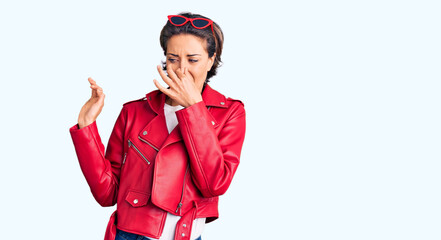 Young beautiful woman wearing red leather jacket smelling something stinky and disgusting, intolerable smell, holding breath with fingers on nose. bad smell
