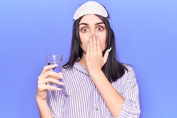 Young beautiful girl wearing sleep mask drinking glass of water covering mouth with hand, shocked...