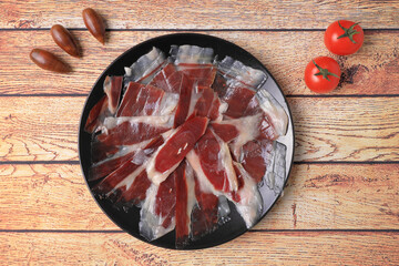 Portion of acorn-fed Iberian ham on black plate on wooden table decorated with acorns and tomatoes