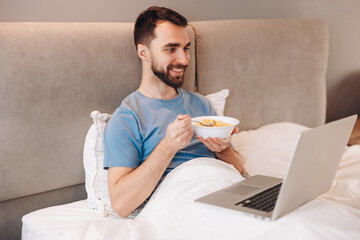 Attractive young man with beard smiling eating breakfast in bed in front of laptop. weekend lazy...
