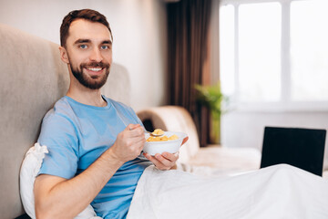 Handsome young man with beard smiling eating breakfast in bed look at camera. weekend lazy day off.