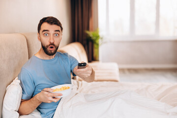 Attractive young man with beard eating breakfast in bed and look disappointed shocked whilst...