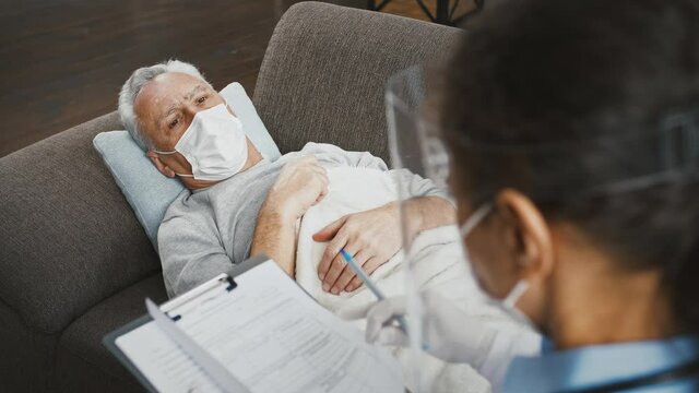 Female doctor in protective mask and face shield discussing symptoms of illness to adjust treatment of aged patient lying on sofa at home