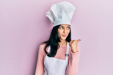 Young hispanic woman wearing baker uniform and cook hat smiling with happy face looking and pointing to the side with thumb up.
