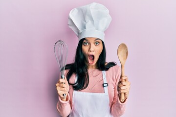 Fototapeta premium Young hispanic woman wearing baker uniform holding spoon and whisk afraid and shocked with surprise and amazed expression, fear and excited face.