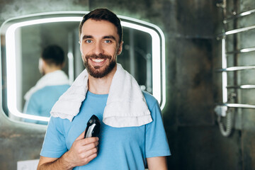 Young smiling man with towel holding brand-new shaver early in the morning at bathroom.