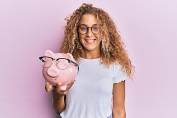 Beautiful caucasian teenager girl holding piggy bank with glasses looking positive and happy...