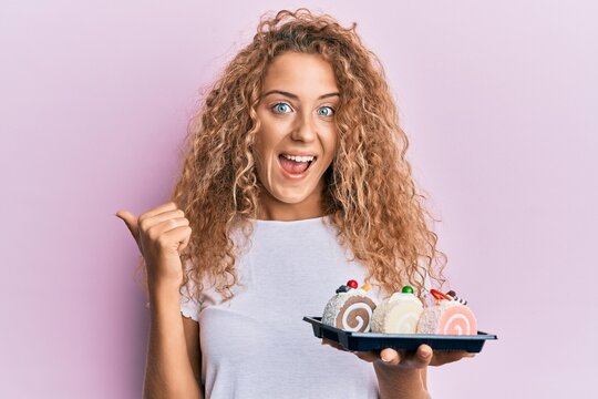 Beautiful caucasian teenager girl holding cake sweets pointing thumb up to the side smiling happy with open mouth