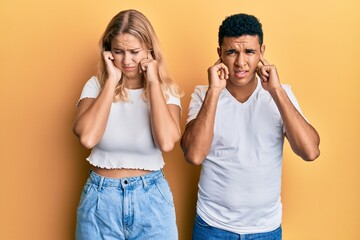 Young interracial couple wearing casual white tshirt covering ears with fingers with annoyed expression for the noise of loud music. deaf concept.