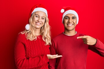 Young interracial couple wearing christmas hat amazed and smiling to the camera while presenting with hand and pointing with finger.