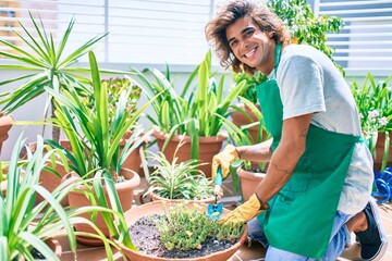 Young hispanic gardener smiling happy caring plants using garden shovel at terrace