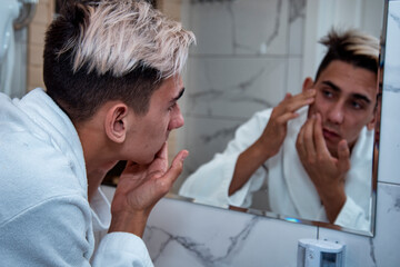 Morning procedures in the bathroom. A young man in a white coat stands at the mirror. Problem skin and care for it. Non-standard appearance.