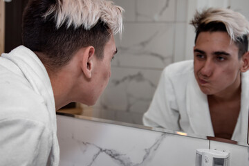 Obraz premium Morning procedures in the bathroom. A young man in a white coat stands at the mirror. Problem skin and care for it. Non-standard appearance.