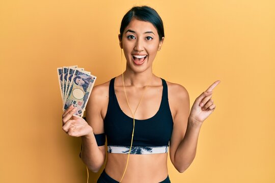 Beautiful asian young sport woman holding 5000 japanese yen banknotes smiling happy pointing with hand and finger to the side