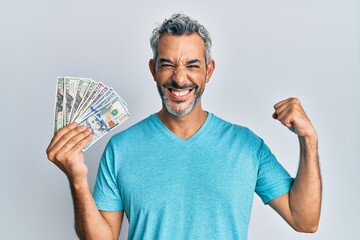 Middle age grey-haired man holding dollars screaming proud, celebrating victory and success very...