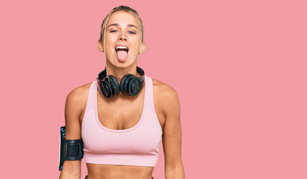 Young blonde woman wearing gym clothes and using headphones sticking tongue out happy with funny expression. emotion concept.