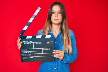 Beautiful caucasian woman holding video film clapboard thinking attitude and sober expression...