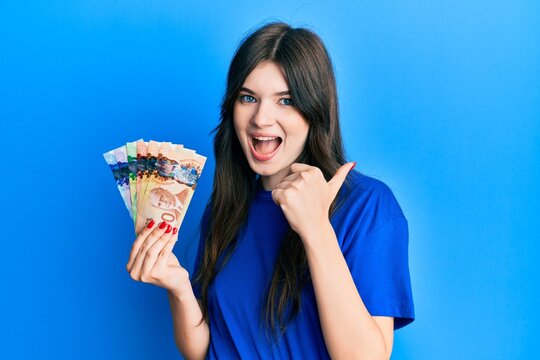 Young beautiful caucasian girl holding canadian dollars pointing thumb up to the side smiling happy with open mouth