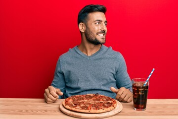 Handsome hispanic man eating tasty pepperoni pizza looking to side, relax profile pose with natural face and confident smile.