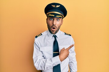 Handsome hispanic man wearing airplane pilot uniform surprised pointing with finger to the side,...