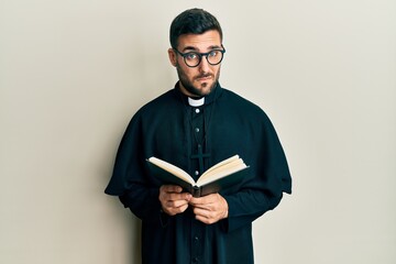 Young hispanic priest man holding bible skeptic and nervous, frowning upset because of problem. negative person. - obrazy, fototapety, plakaty