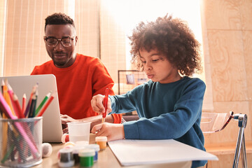 Boy drawing near his father working with laptop at kitchen