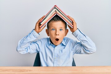 Adorable caucasian kid holding book on head afraid and shocked with surprise and amazed expression,...