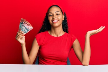 Young african american girl holding australian dollars sitting on the table celebrating achievement with happy smile and winner expression with raised hand