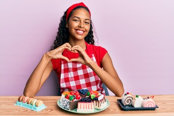 Young african american girl wearing baker uniform sitting on the table with sweets smiling in love doing heart symbol shape with hands. romantic concept.