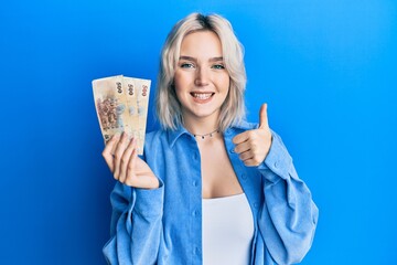 Young blonde girl holding new taiwan dollars banknotes smiling happy and positive, thumb up doing excellent and approval sign