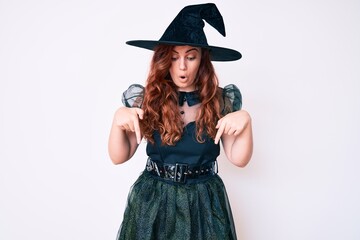 Young beautiful woman wearing witch halloween costume pointing down with fingers showing advertisement, surprised face and open mouth