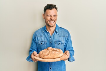 Young hispanic man holding bread winking looking at the camera with sexy expression, cheerful and...