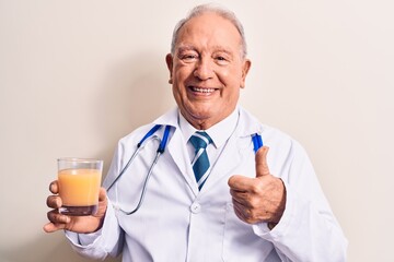 Senior grey-haired doctor man wearing stethoscope drinking glass of healthy orange juice smiling...