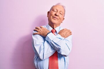 Senior handsome grey-haired businessman wearing elegant tie standing over pink background hugging oneself happy and positive, smiling confident. Self love and self care