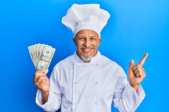 Middle age grey-haired man wearing professional cook uniform holding usa dollars banknotes smiling happy pointing with hand and finger to the side