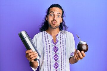 Young handsome man with long hair drinking mate infusion puffing cheeks with funny face. mouth...