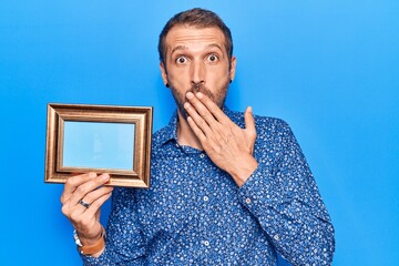 Young handsome man holding empty frame covering mouth with hand, shocked and afraid for mistake....